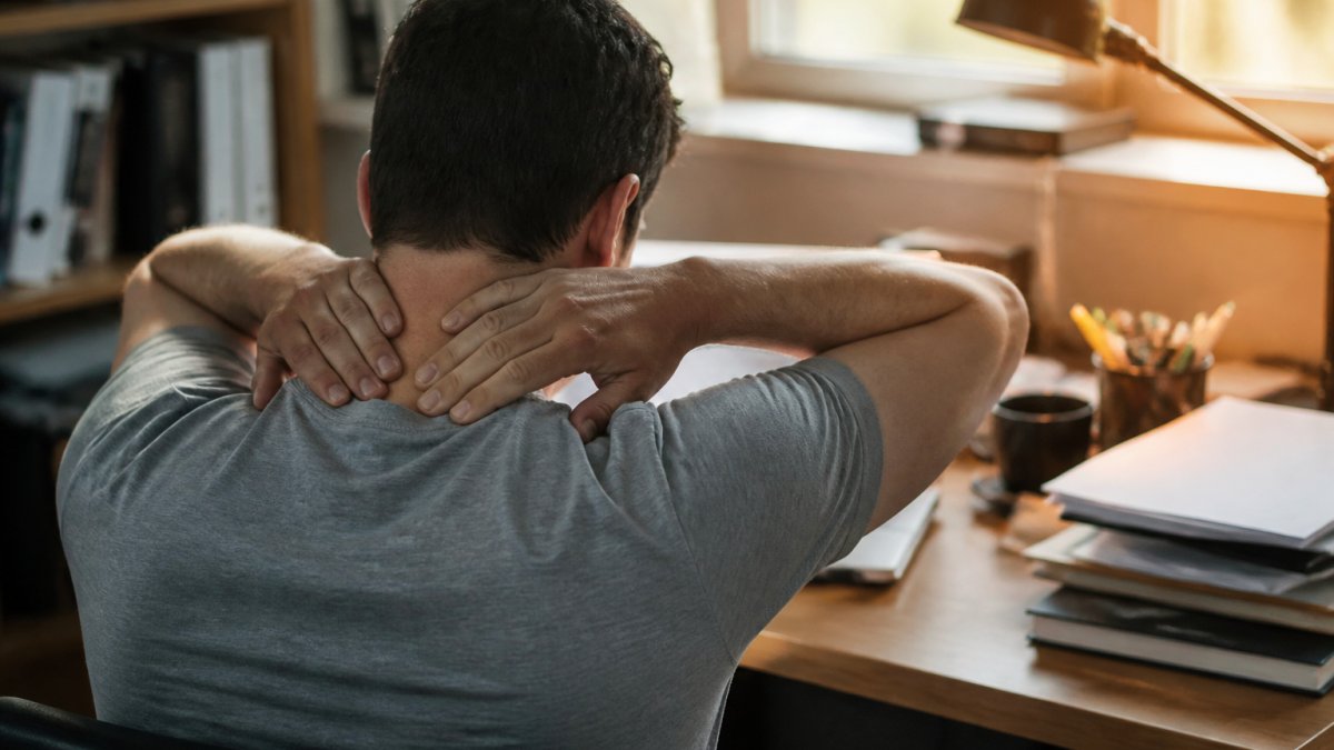 A close-up of a person's hands massaging the base of their own neck while sitting at a cluttered office desk, warm afternoon light through a window
